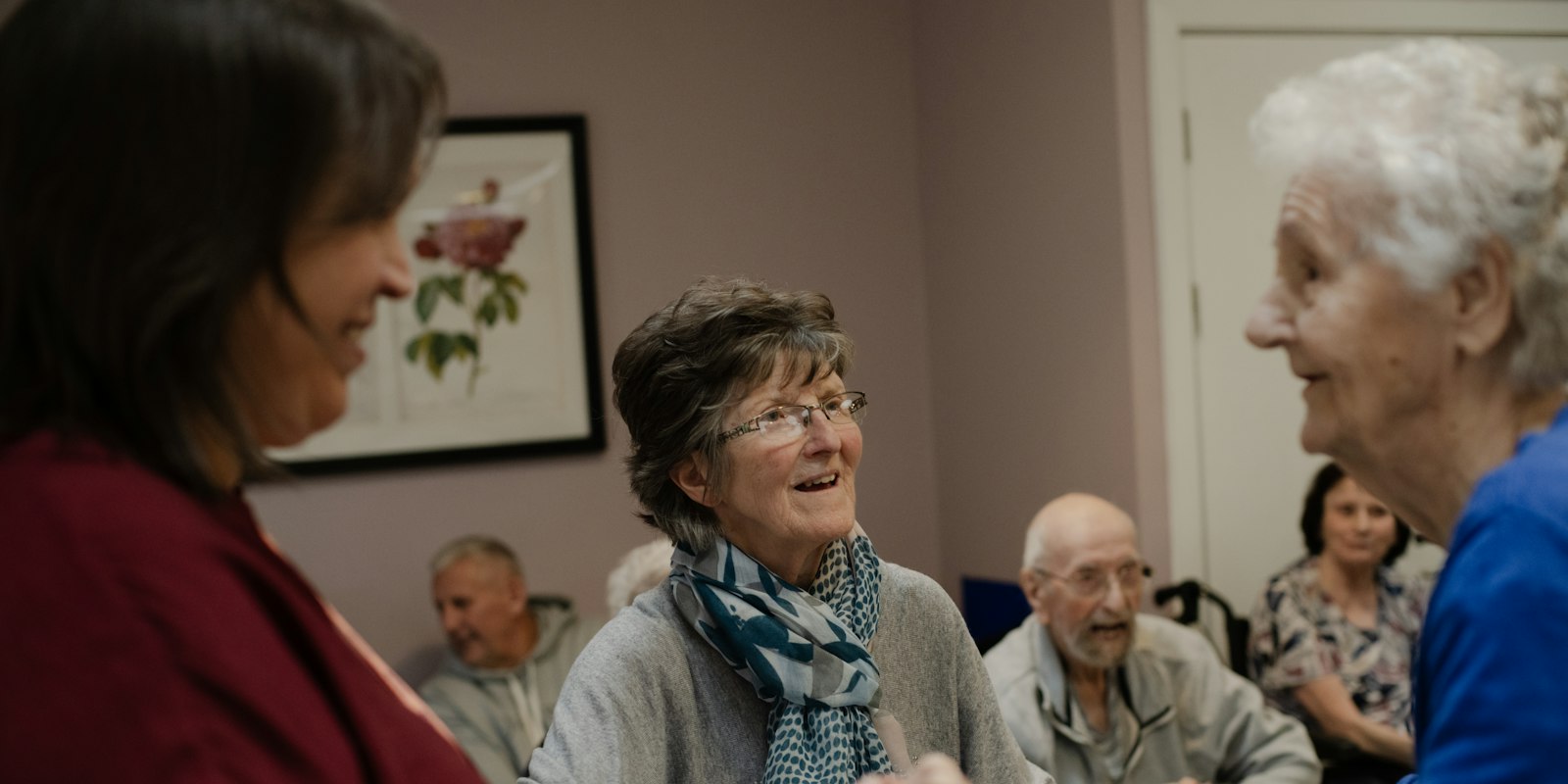 Drie vrouwen lachen samen in een huiskamer, met oudere mensen op de achtergrond.